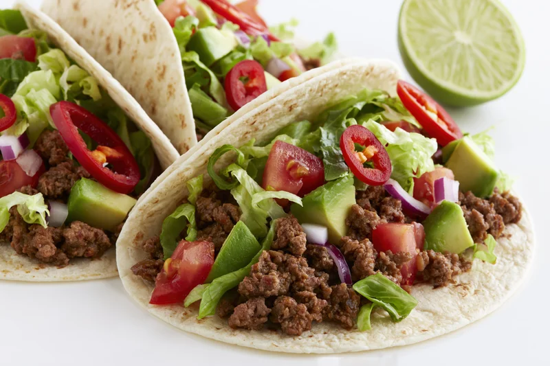 Two flour tortilla tacos filled with seasoned ground beef and topped with lettuce, tomato, avocado, red onion, and sliced chili, with a lime wedge on the side, on a white surface