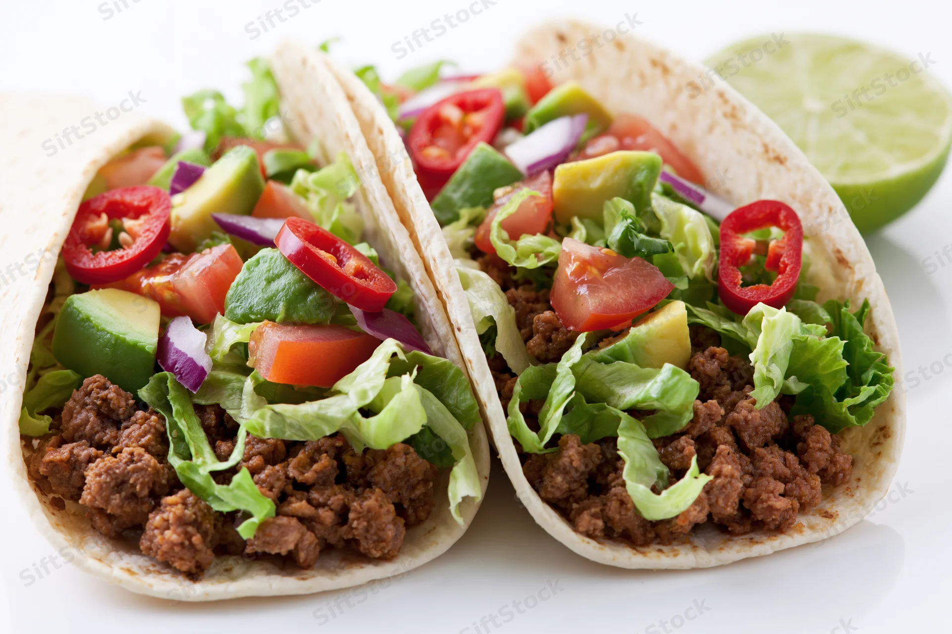 Two flour tortilla tacos filled with seasoned ground beef and topped with lettuce, tomato, avocado, red onion, and chili slices, served with a lime wedge on a white surface