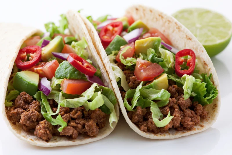 Two flour tortilla tacos filled with seasoned ground beef and topped with lettuce, tomato, avocado, red onion, and chili slices, served with a lime wedge on a white surface
