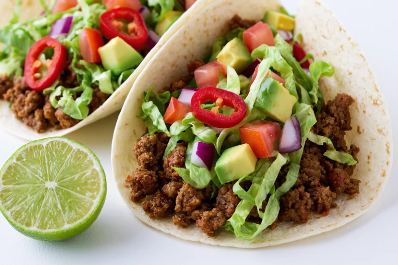 Two tacos filled with seasoned ground beef and fresh toppings arranged on a white surface with a lime wedge beside them
