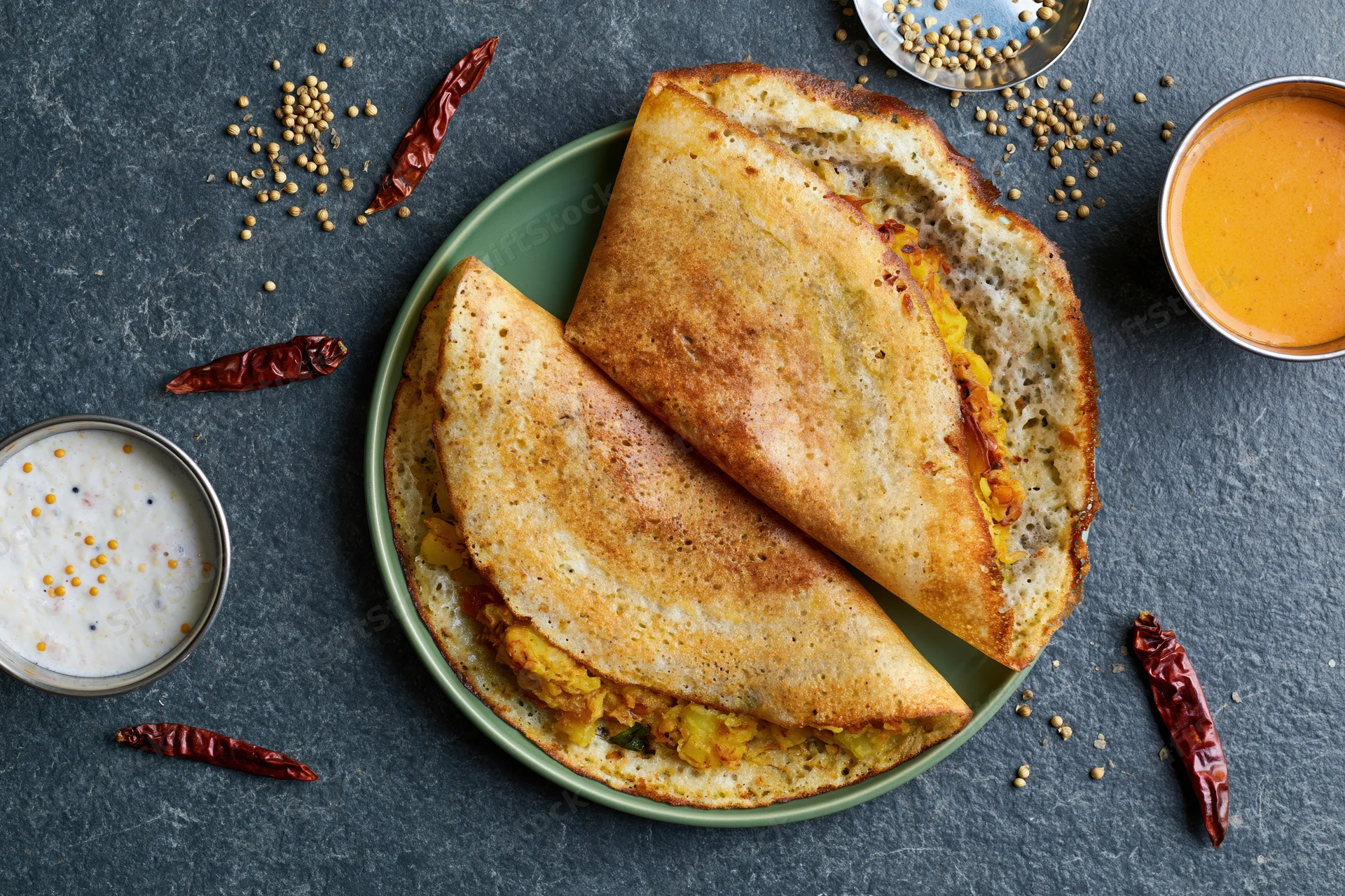 Two folded masala dosas with golden-brown edges and potato filling on a green plate with chutney and sambar on a dark slate surface