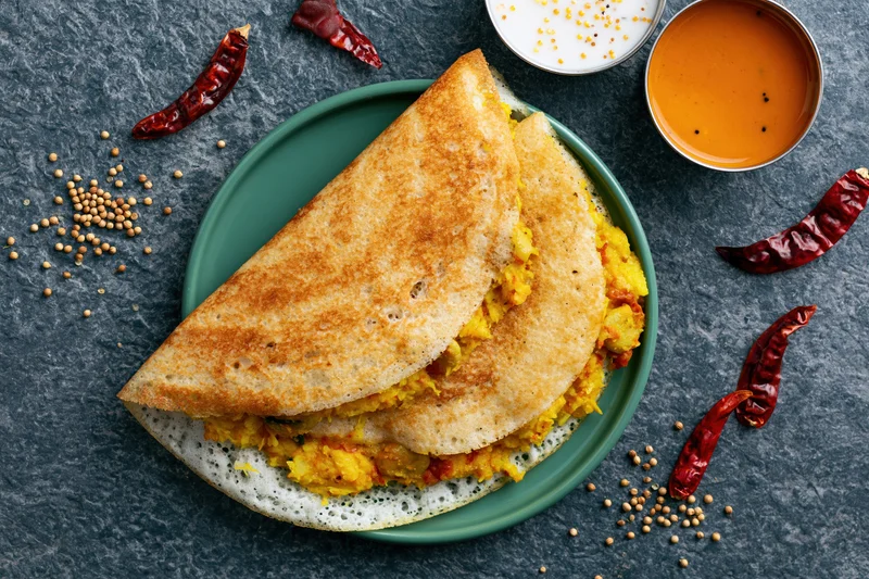 Two folded masala dosas with golden-brown edges and yellow filling on a green plate with chutney, dried chilies, and mustard seeds on a dark stone surface