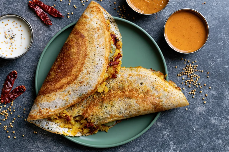Two folded masala dosas with golden-brown crisp edges and spiced potato filling on a green plate with chutney and sambar bowls, surrounded by spices on a dark stone surface