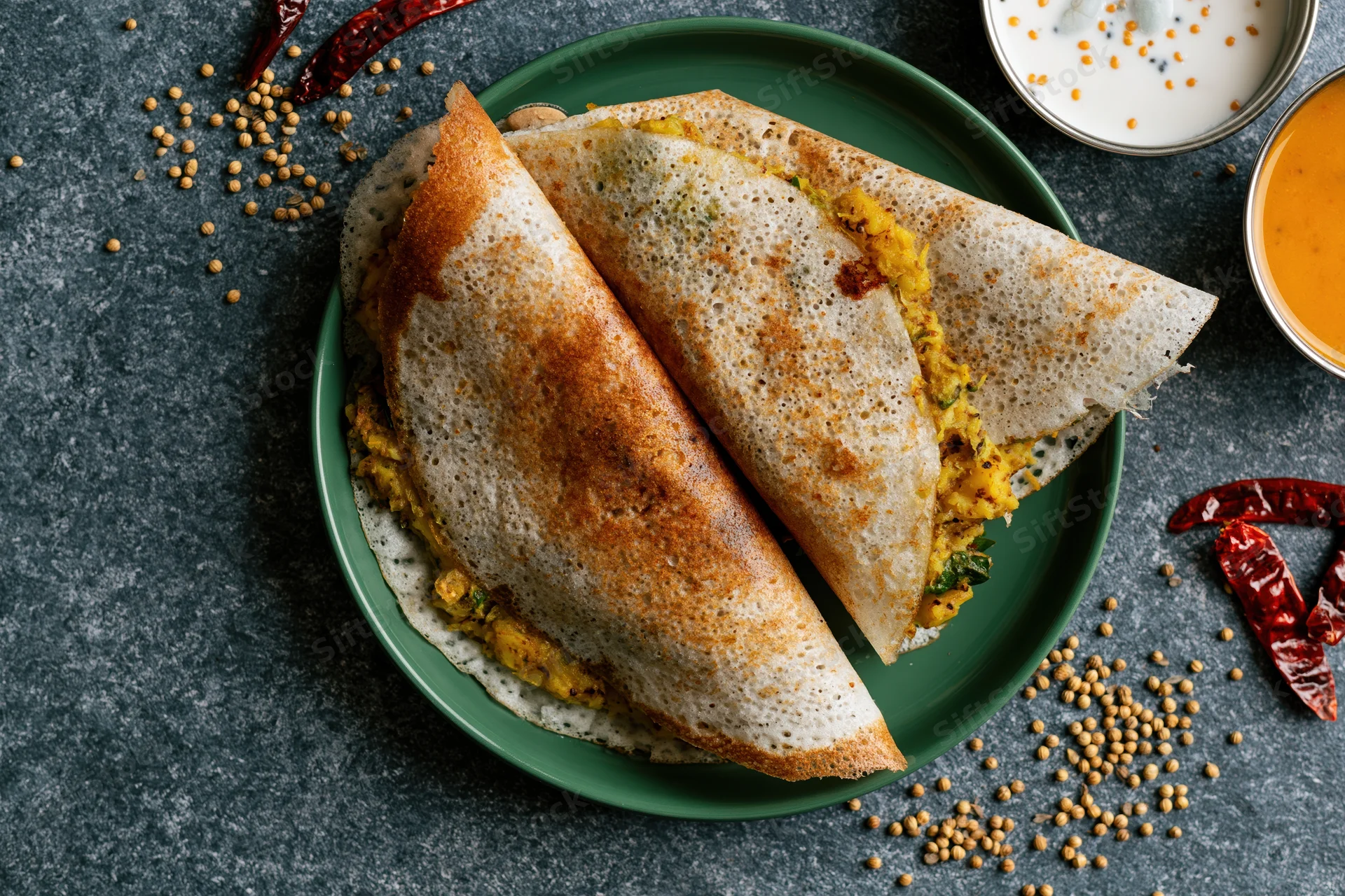 Two folded masala dosas with golden-brown crisp edges and spiced potato filling on a green plate with chutney and spices around