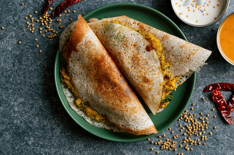 Two folded masala dosas with golden-brown crisp edges and spiced potato filling on a green plate with chutney and spices around