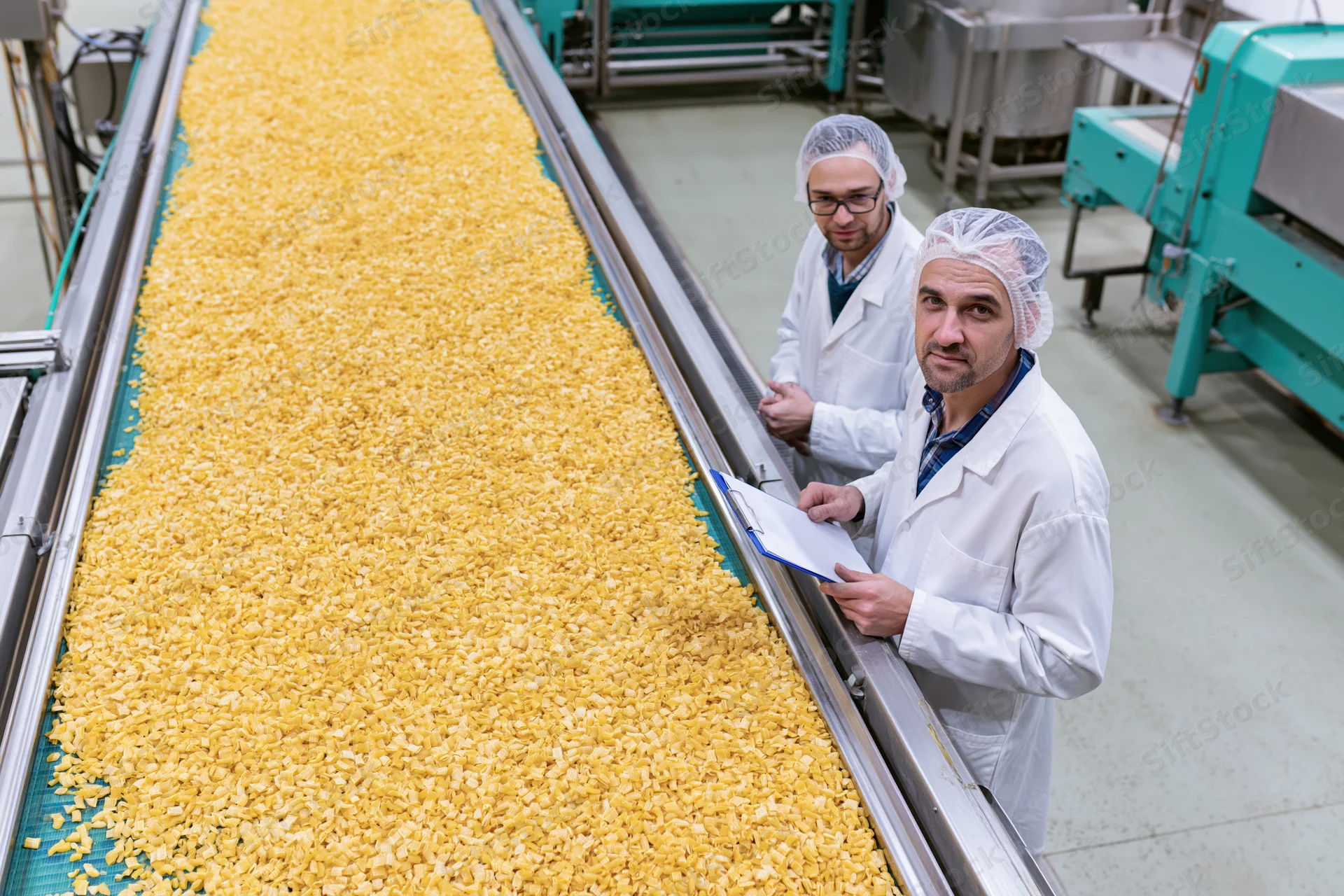 Two men in lab coats and hairnets inspecting pasta on a conveyor belt in a food factory