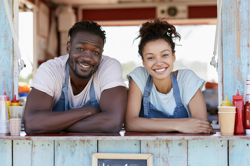 Young man and woman in aprons smiling while leaning on food truck counter