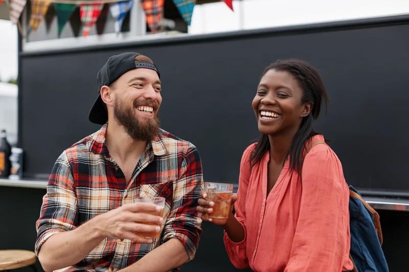 Two friends laughing and holding drinks while seated on stool outdoors at a festival