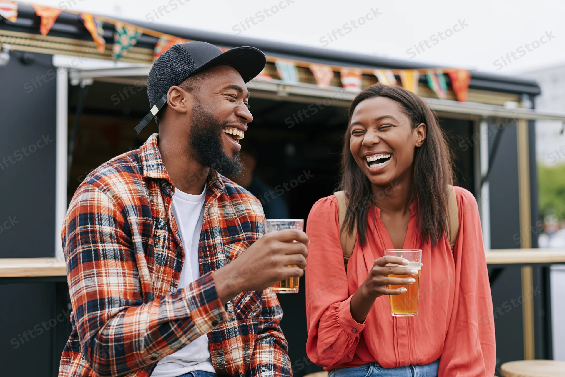 Two friends laughing and holding beer glasses while seated at a food truck outdoor event