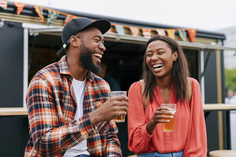 Two friends laughing and holding beer glasses while seated at a food truck outdoor event