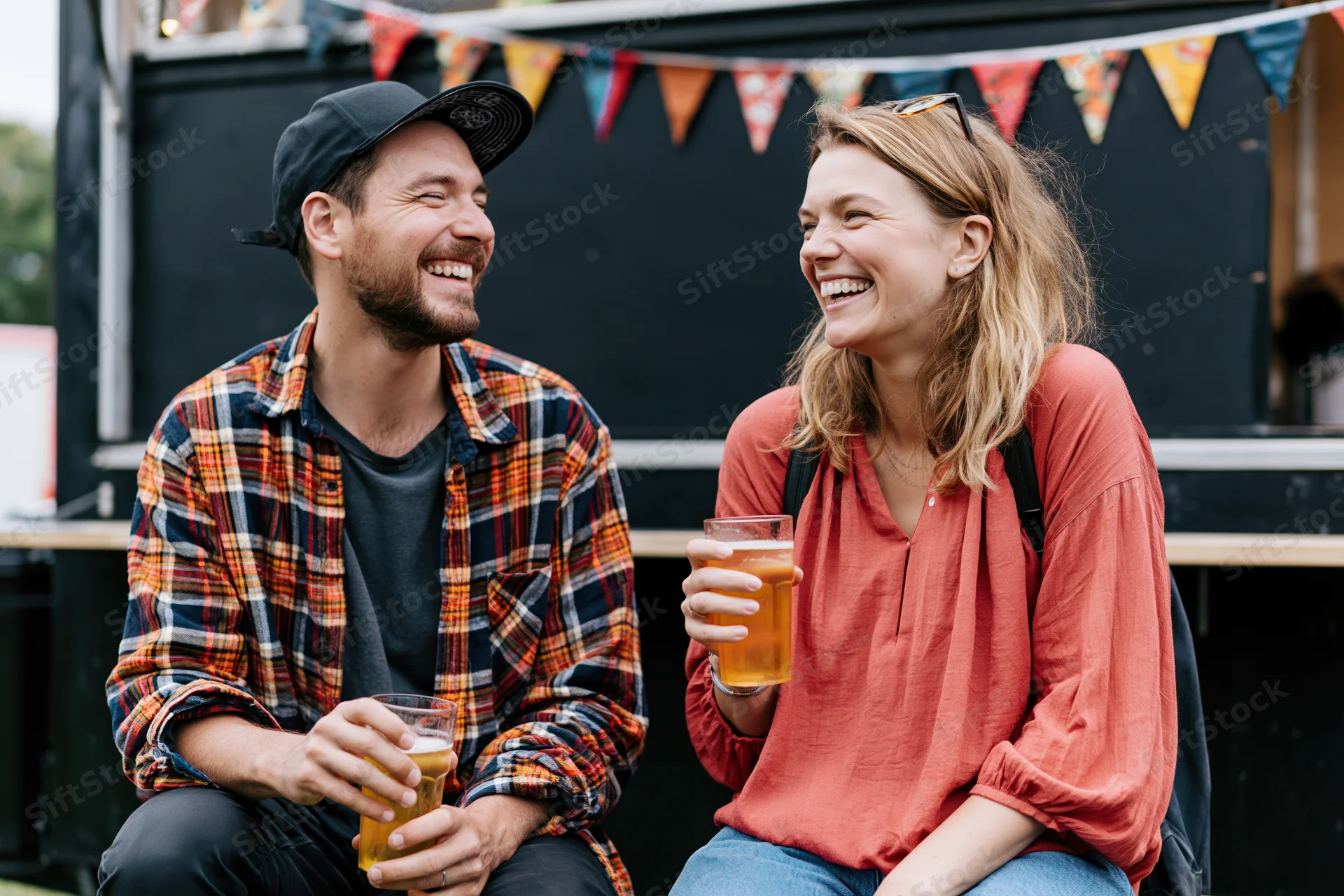 Two friends laughing and holding drinks while seated outdoors at a festival