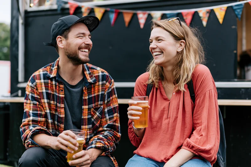 Two friends laughing and holding drinks while seated outdoors at a festival