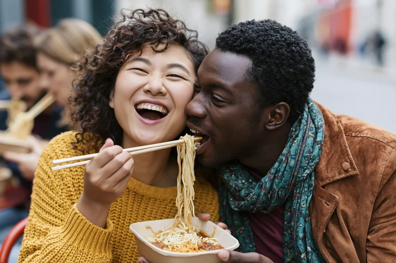 Couple laughing and sharing noodles from a takeout container outdoors