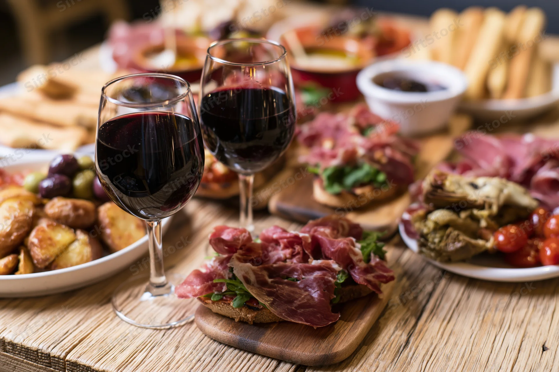 Two glasses of red wine on a rustic wooden table with charcuterie and appetizers arranged around them
