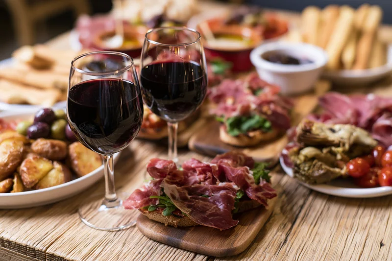 Two glasses of red wine on a rustic wooden table with charcuterie and appetizers arranged around them