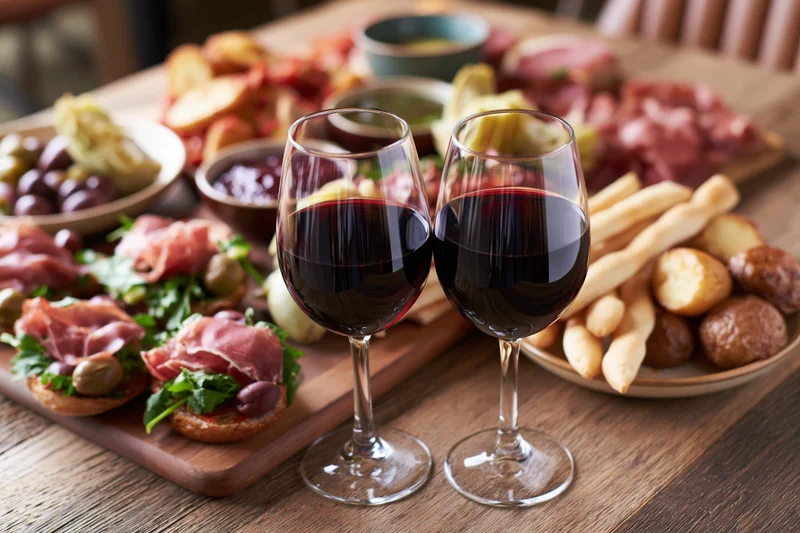 Two glasses of red wine on a wooden table with charcuterie board and appetizers