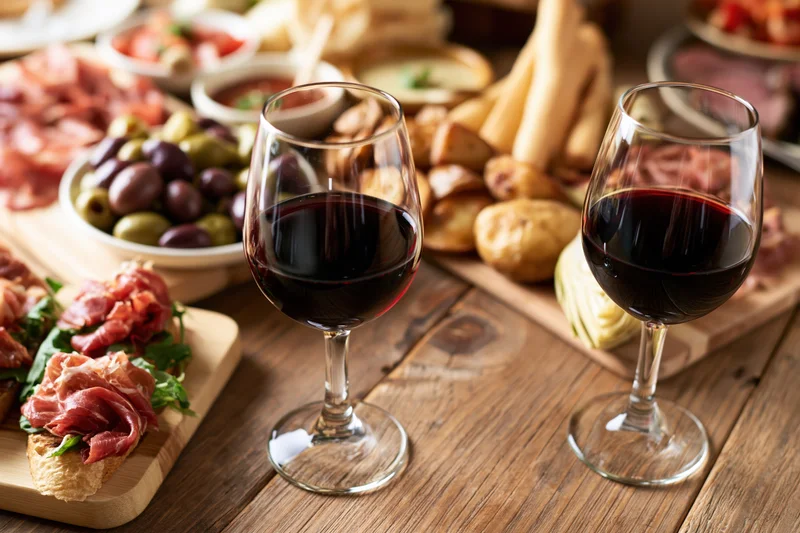Two glasses of red wine on a rustic wooden table surrounded by charcuterie and appetizers