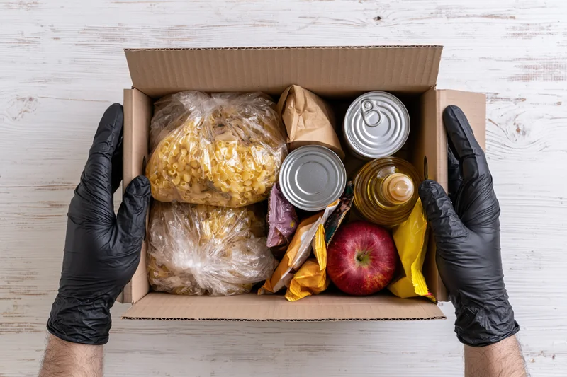 Gloved hands holding open cardboard box filled with groceries on white wooden surface