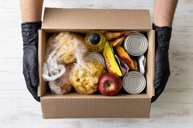 Gloved hands holding open cardboard box filled with food staples and groceries on white wooden surface