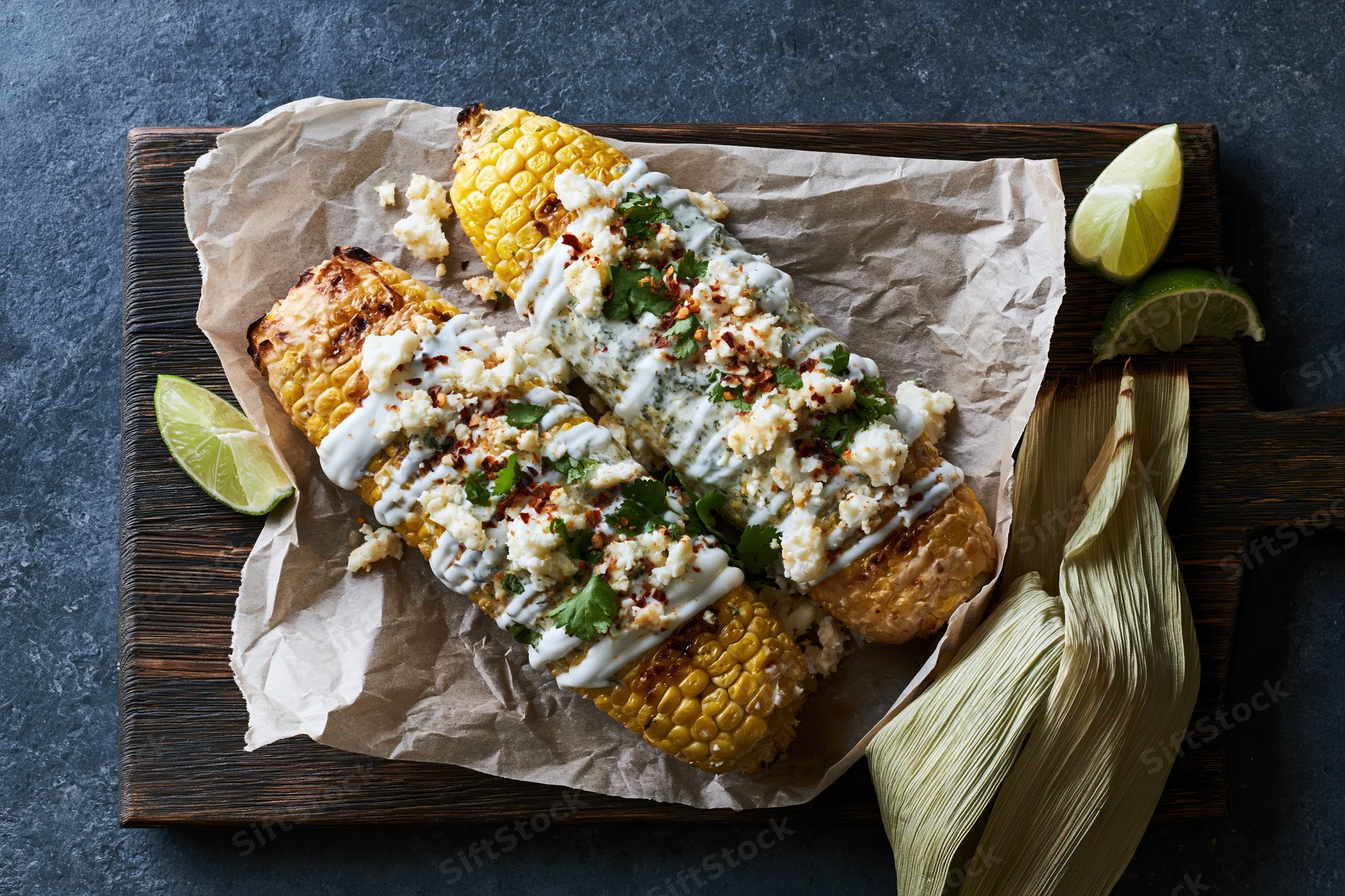 Grilled corn cobs topped with cotija cheese, lime, and cilantro on parchment paper on a wooden board
