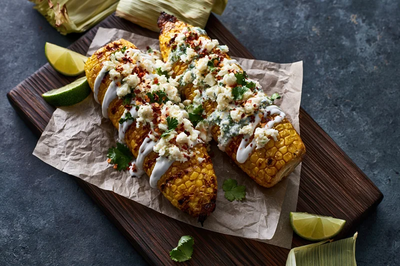 Grilled corn cobs topped with crumbled cotija cheese, lime, and cilantro on a wooden cutting board