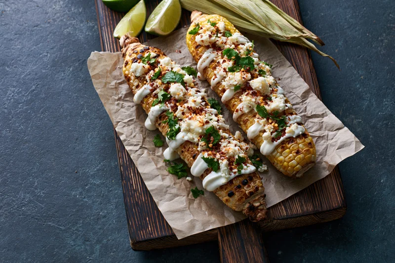 Grilled corn cobs topped with cotija cheese, cilantro, lime, and sauce on parchment paper on a wooden cutting board