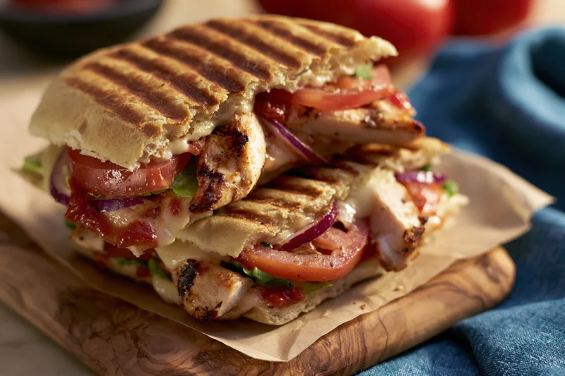 Two grilled chicken panini sandwiches stacked on a wooden olive board