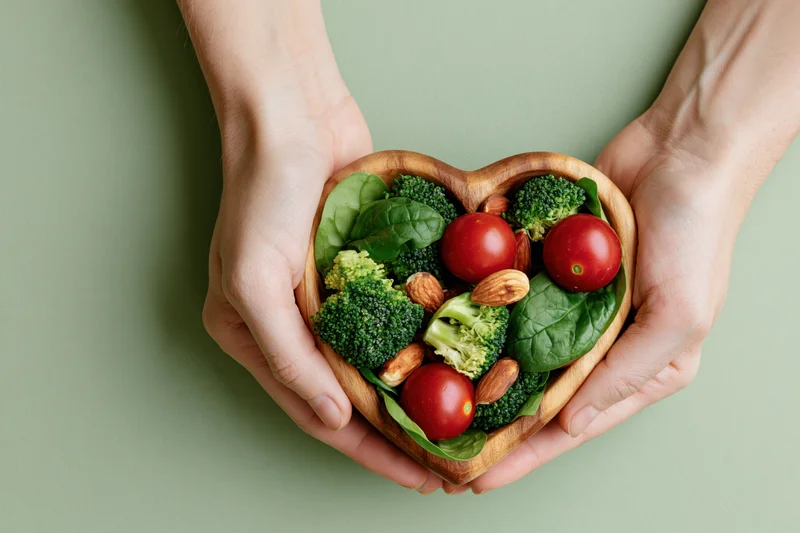 Hands holding a heart-shaped wooden bowl filled with broccoli, tomatoes, spinach and almonds against a green background
