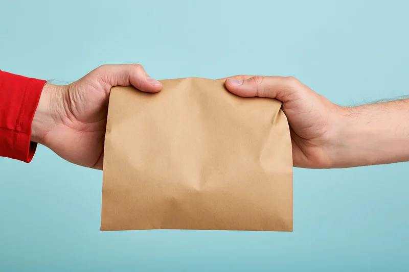 Two hands exchanging a plain kraft paper bag against a light blue background