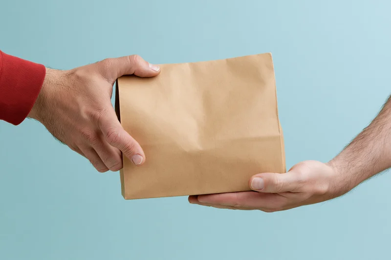 Two hands exchanging a brown kraft paper bag against a light blue background