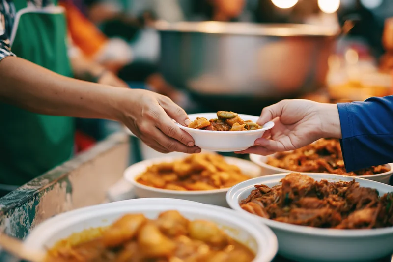 Hands exchanging a small white bowl of stewed food across a table at an outdoor charity event