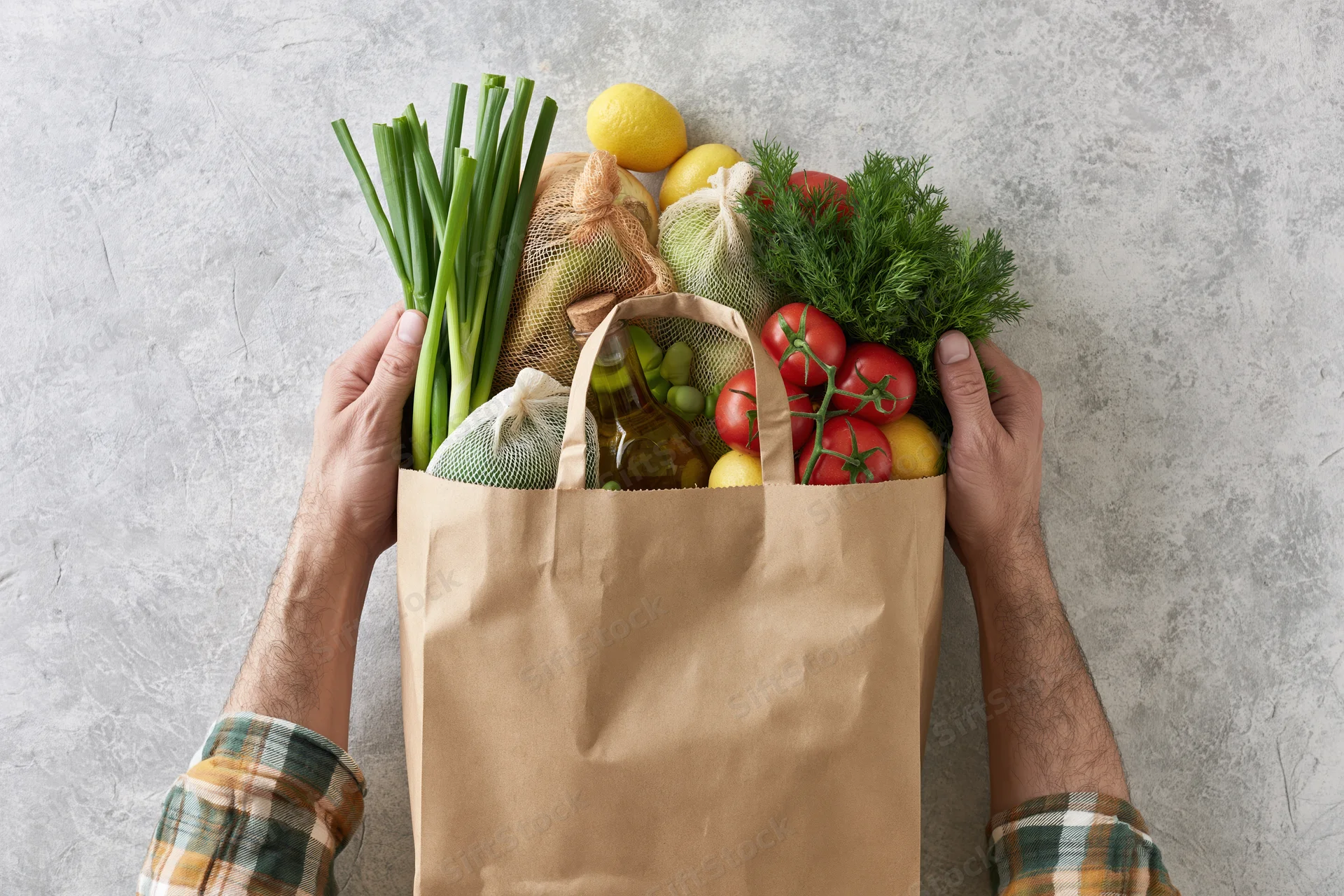 Man holding a brown paper grocery bag filled with fresh vegetables and herbs on a gray countertop