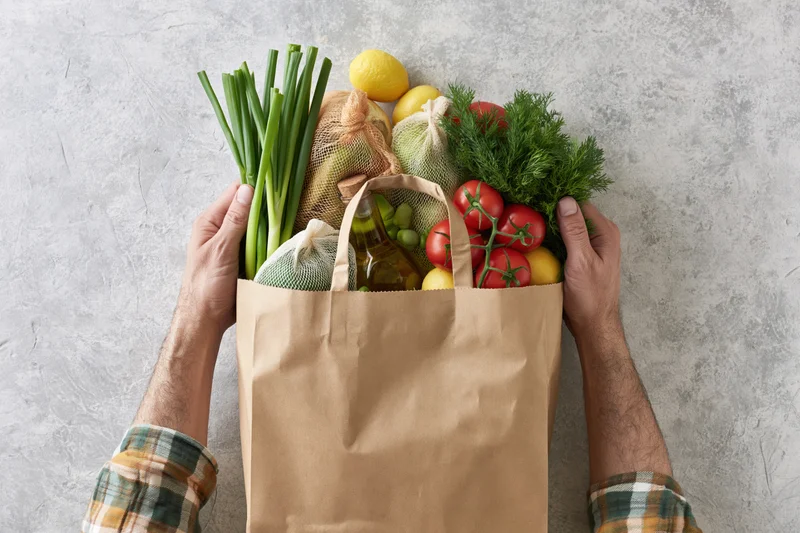 Man holding a brown paper grocery bag filled with fresh vegetables and herbs on a gray countertop