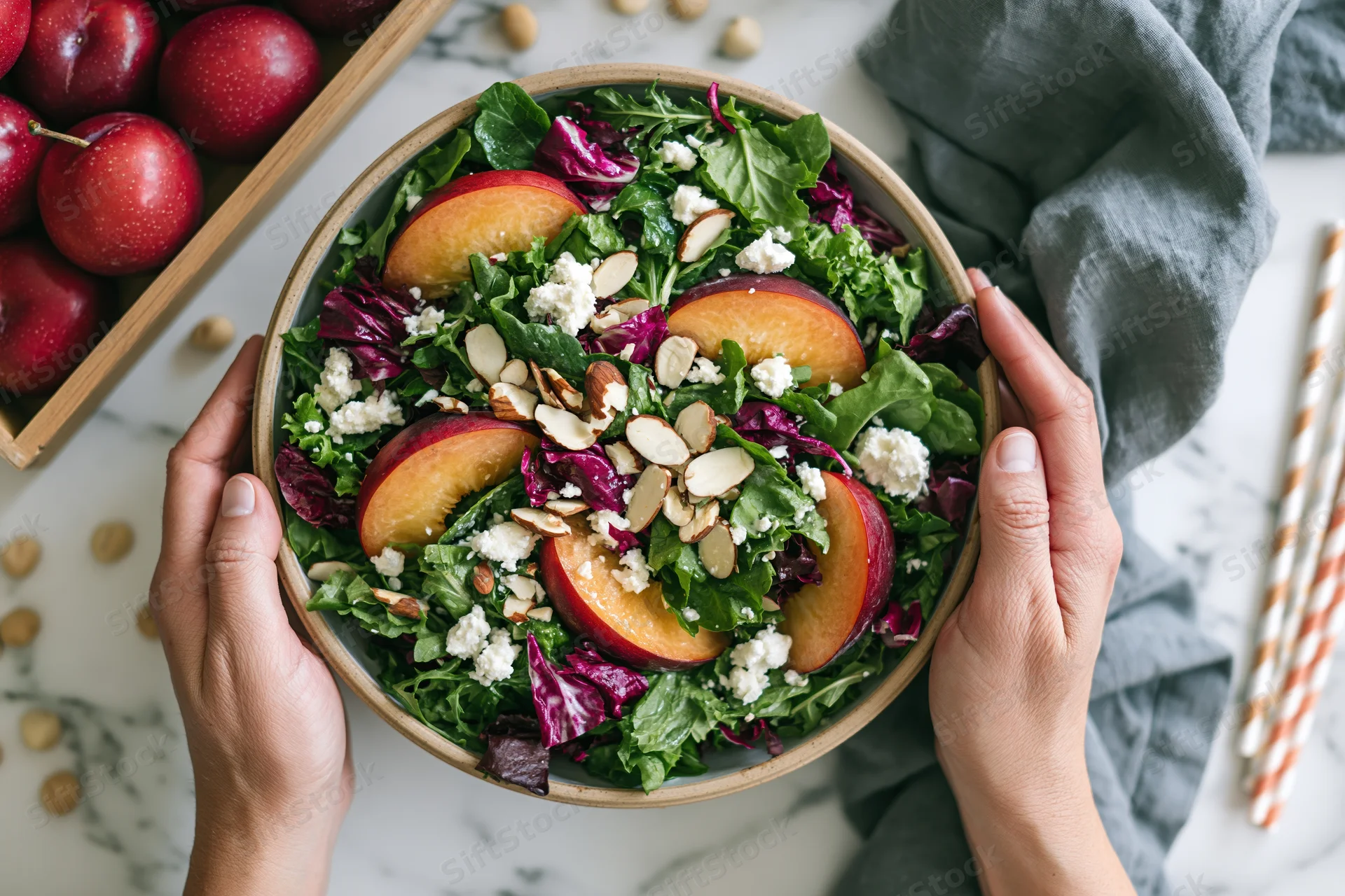 Hands holding a ceramic bowl of stone fruit salad with greens and feta on a marble countertop