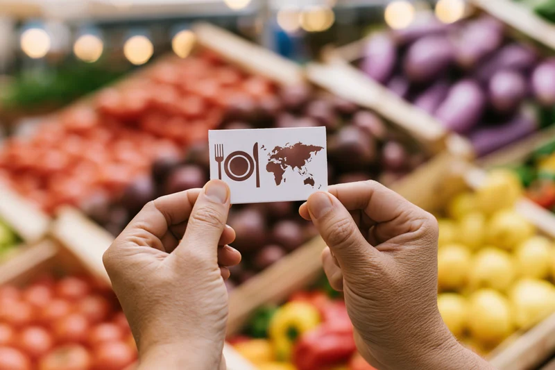 Hands holding a card with a plate and world map icon in front of a vegetable market display