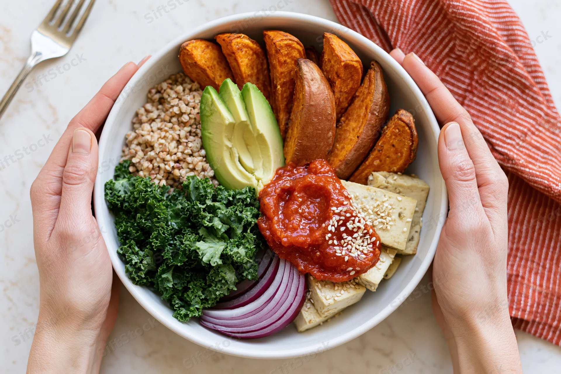 Hands holding a white ceramic bowl filled with vegan buddha bowl ingredients including sweet potato, avocado, kale, red onion, tofu, sauce, and grains on a kitchen countertop