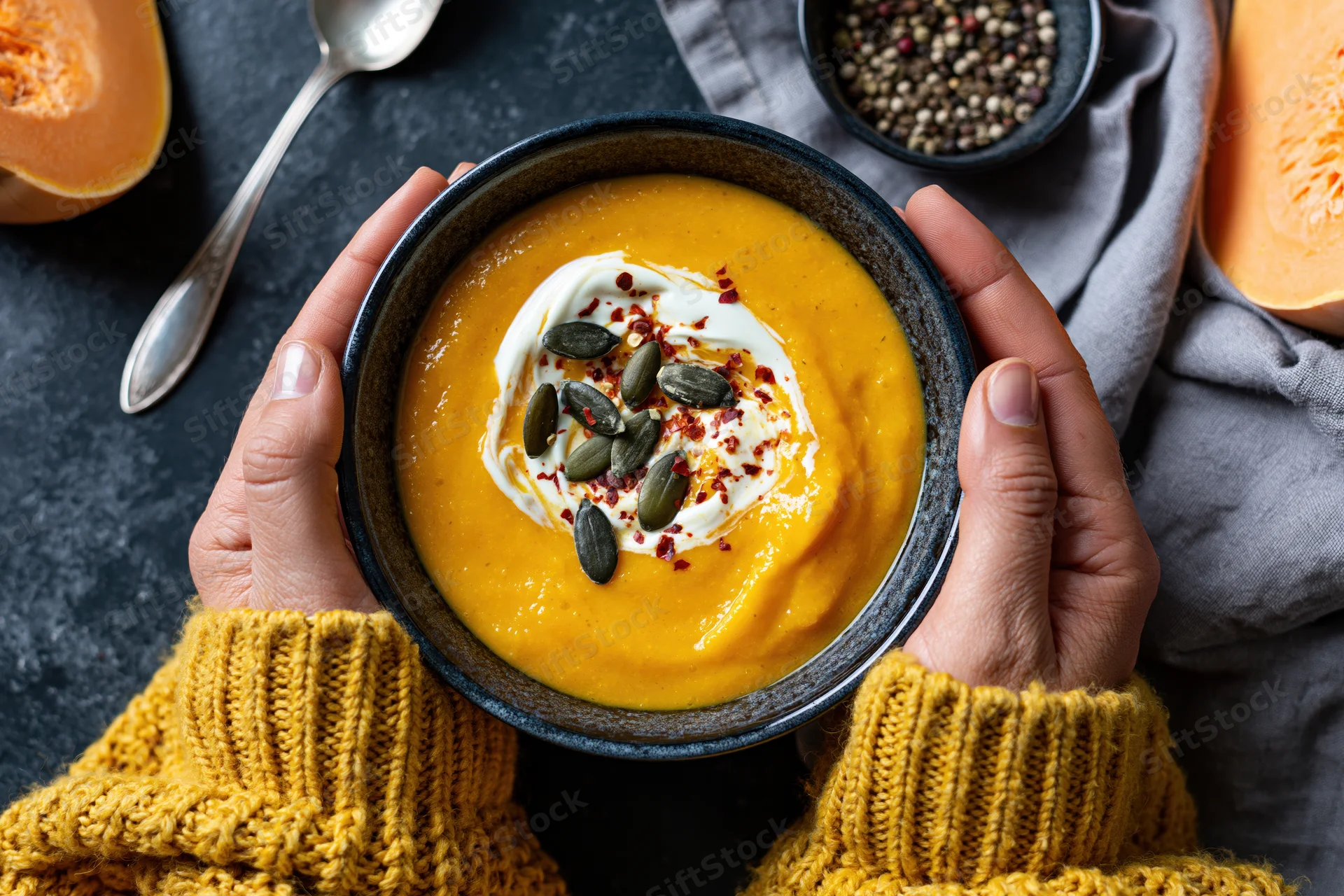 Hands in a yellow knit sweater holding a bowl of pumpkin soup with cream and seeds on a dark table