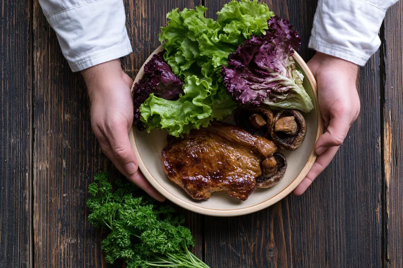 Hands holding a ceramic plate with roasted chicken, mushrooms, and mixed greens on a dark wooden table