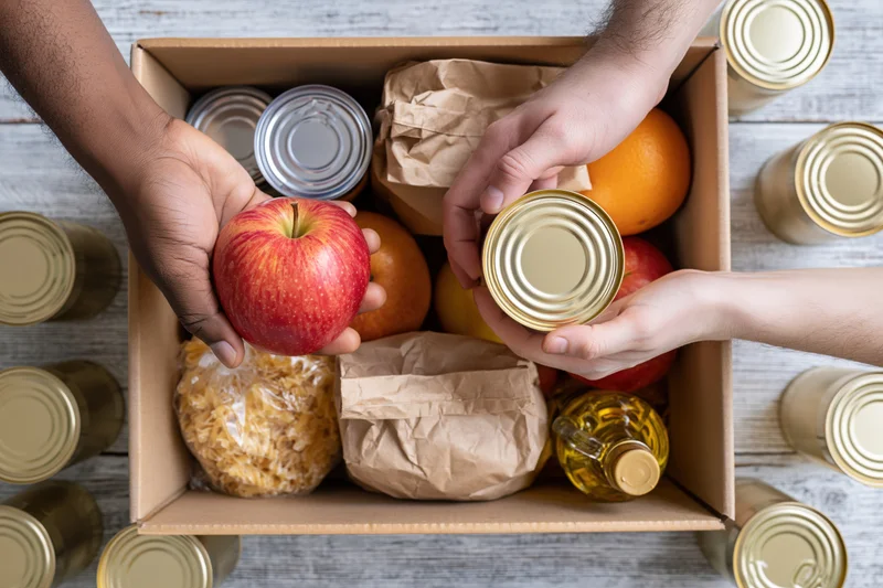 Two hands placing an apple and a can into a food donation box on a wooden table