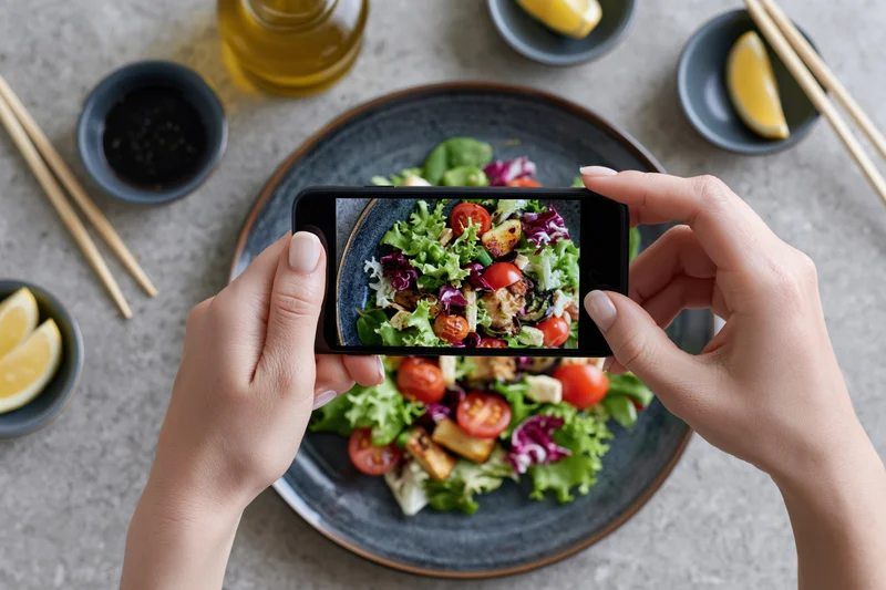 Hands holding smartphone horizontally to photograph salad on plate with chopsticks and lemon wedges on table