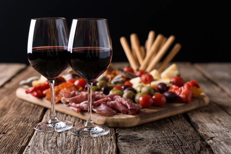 Two wine glasses filled with red wine in front of a charcuterie board on a rustic wooden table
