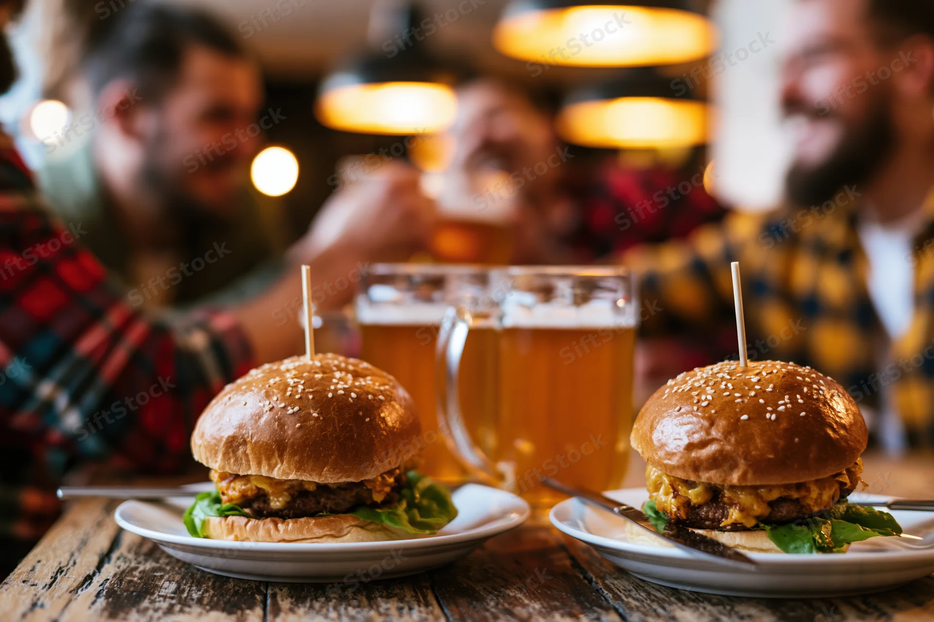 Two loaded burgers on white plates with beer mugs on a wooden table in a pub with men in plaid shirts laughing in the background
