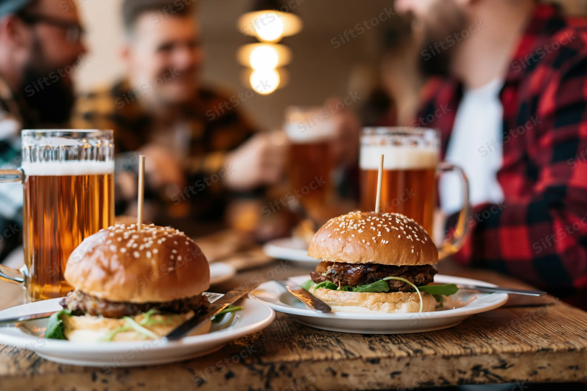 Two loaded pub burgers on white plates with beer mugs on a wooden table, surrounded by men socializing in a warm-lit restaurant interior