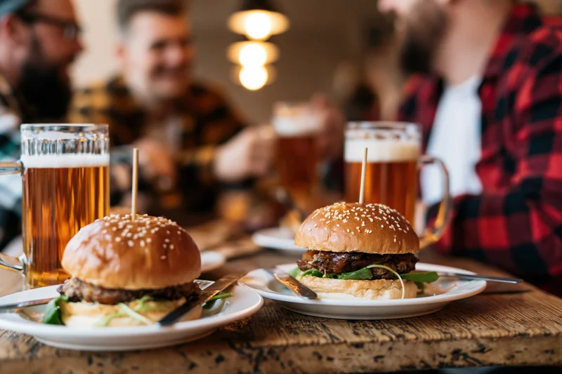 Two loaded pub burgers on white plates with beer mugs on a wooden table, surrounded by men socializing in a warm-lit restaurant interior