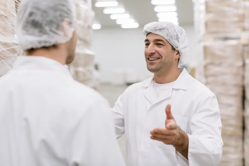 Two male food factory workers in white lab coats and hairnets talking in a warehouse setting
