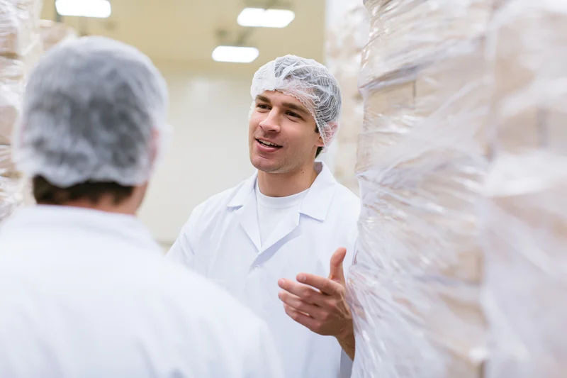 Two male workers in white lab coats and hairnets discussing in a food factory warehouse