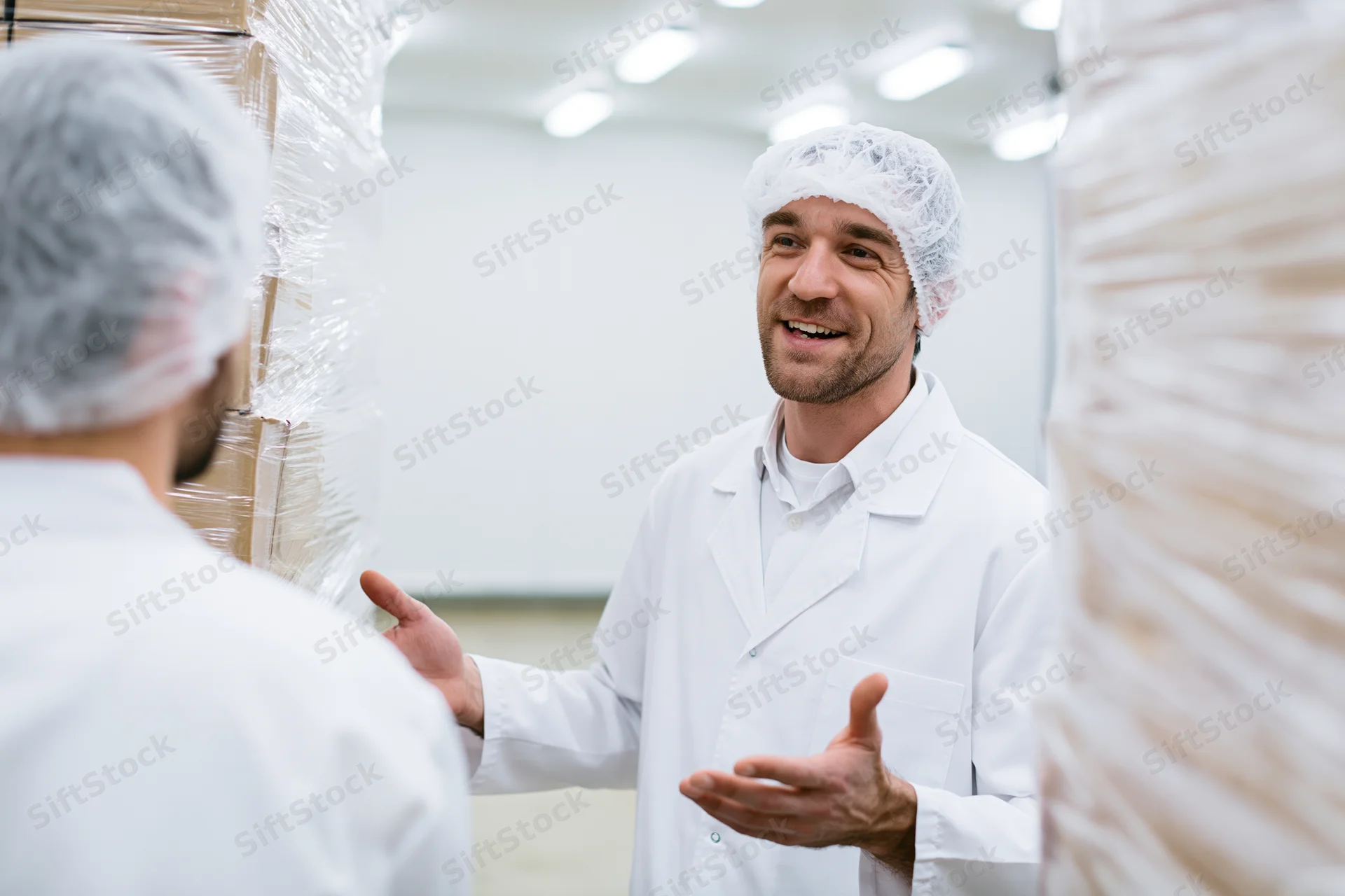 Male worker in white lab coat and hairnet talking to colleague in food factory aisle