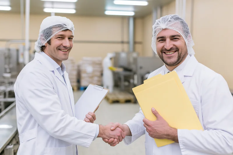 Two men in lab coats and hairnets shaking hands in a food factory interior
