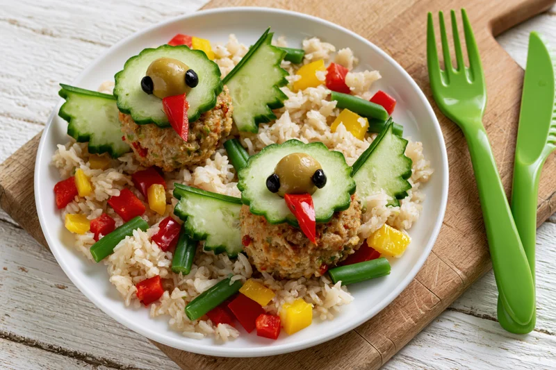 Two meatball chicks arranged as playful bird shapes on a white plate with rice and vegetables, served on a wooden board with green cutlery