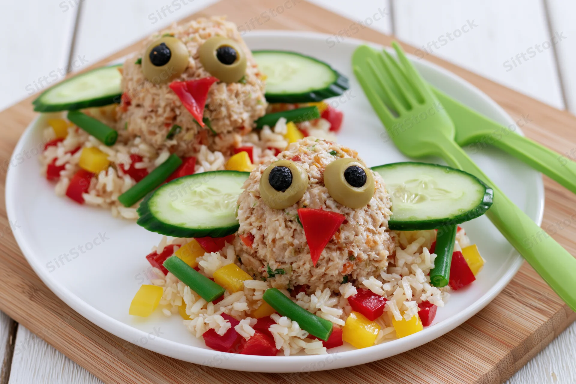 Two meatball chicks shaped like birds on a plate with rice and vegetables, served with green plastic cutlery on a wooden board